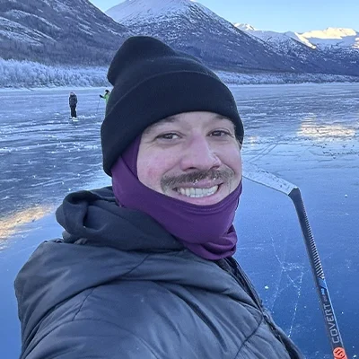 Outdoor photo of Iain McKeever standing on a frozen lake, wearing winter clothing and holding a hockey stick.